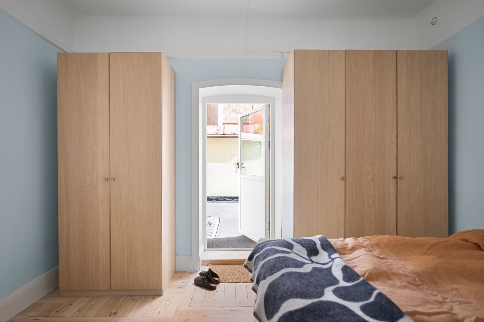 Bedroom with two tall wooden wardrobes standing flush against pale blue walls either side of an open external door