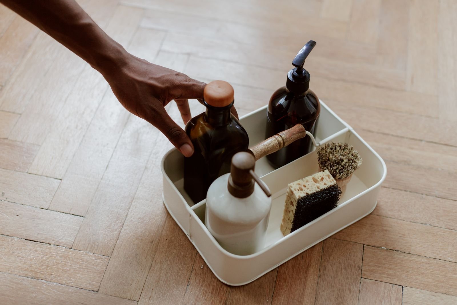 Hand reaching for a cleaning caddy holding soap dispensers, a scrubbing brush and a sponge on a wooden floor