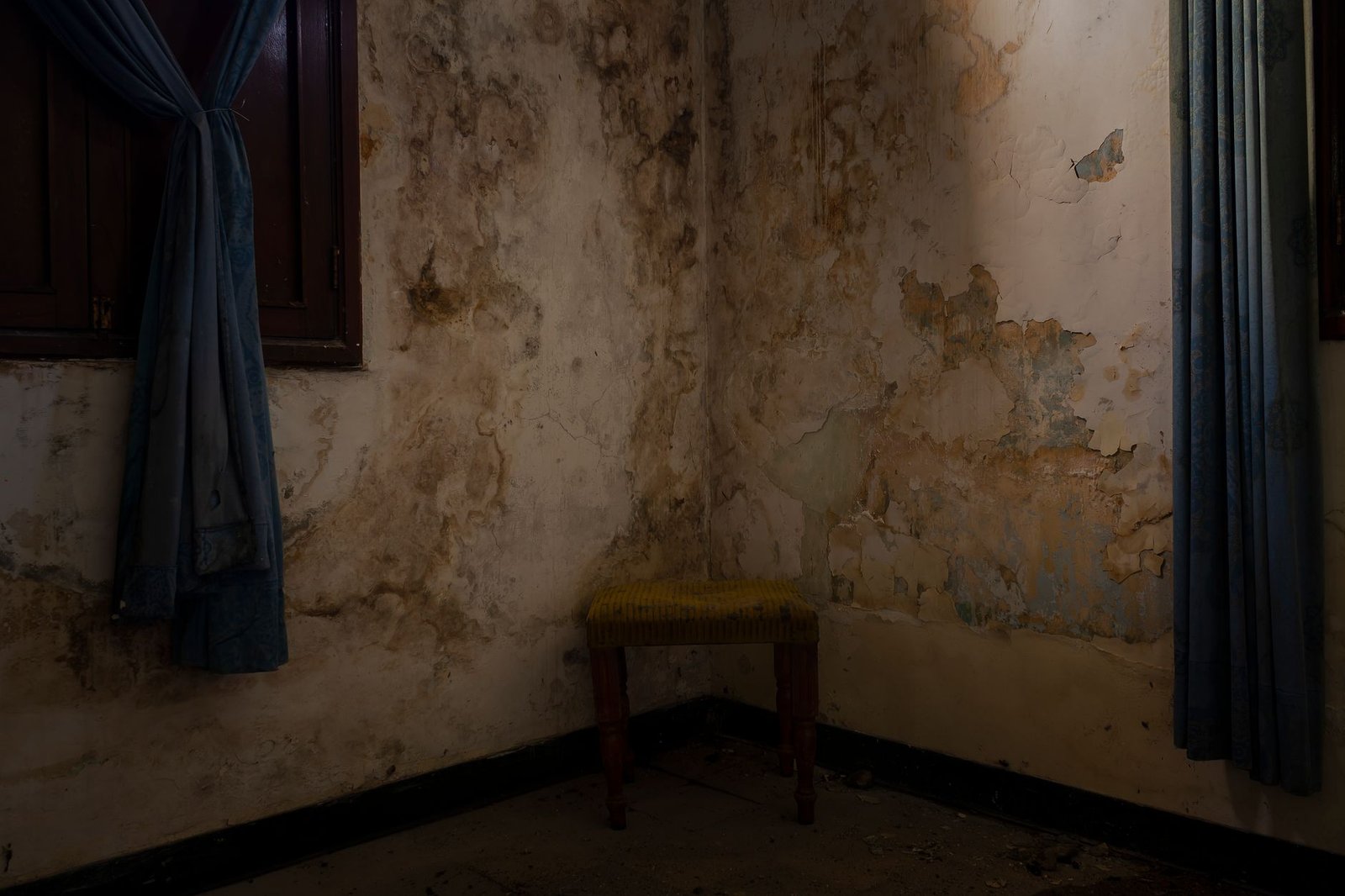 Damp-stained, peeling plaster walls in the corner of a disused room beside a window