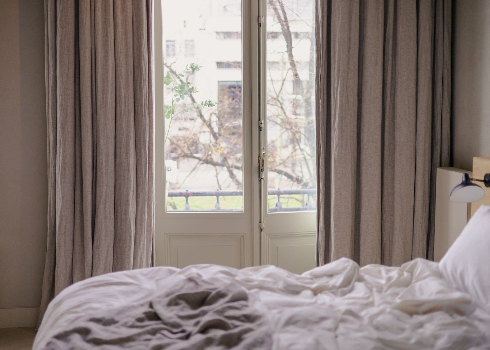 Floor-length linen curtains hanging either side of a white-framed French window above an unmade bed