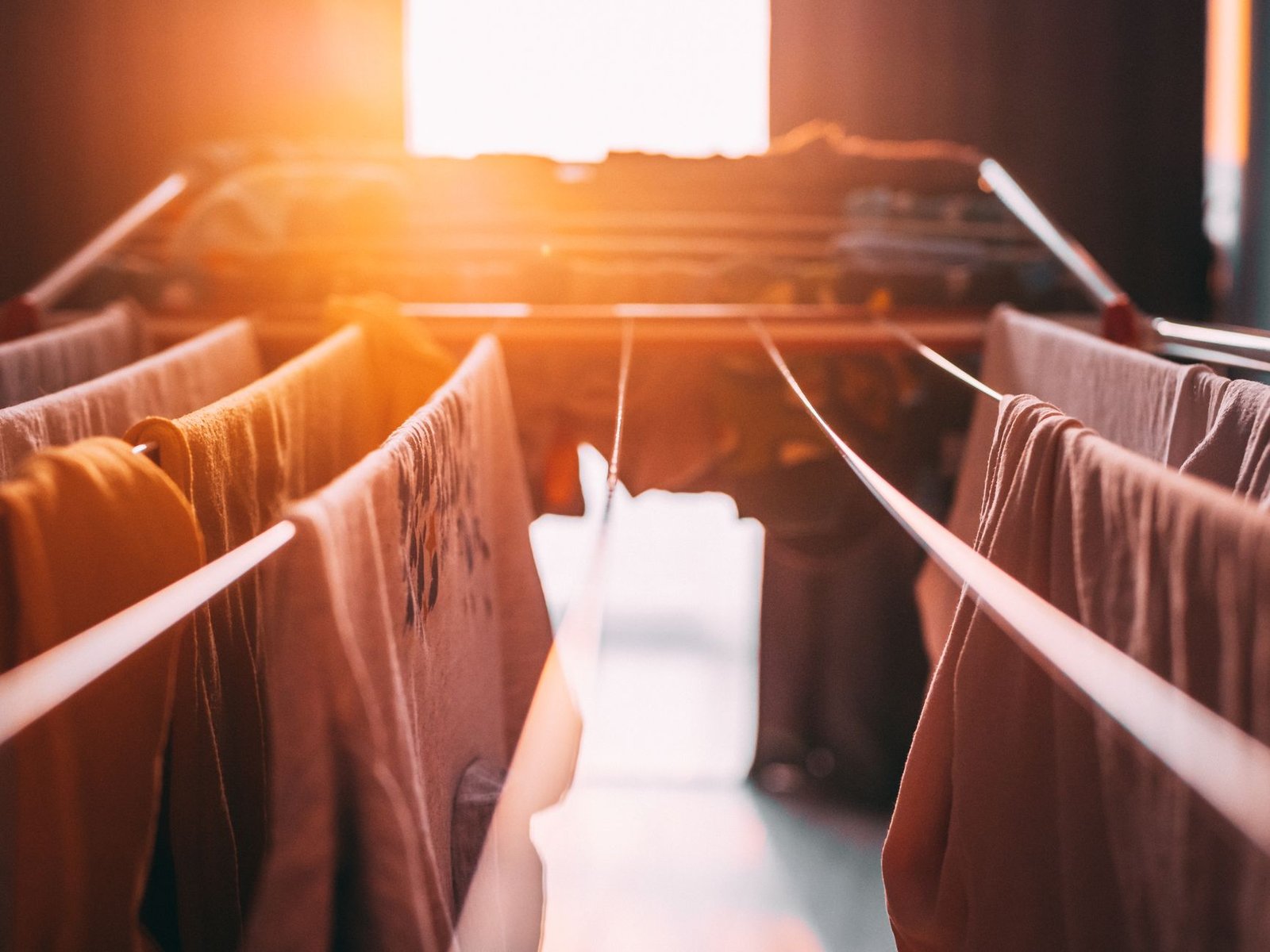 Garments hung apart on the bars of an indoor drying rack in low sunlight, with gaps for air to pass between them