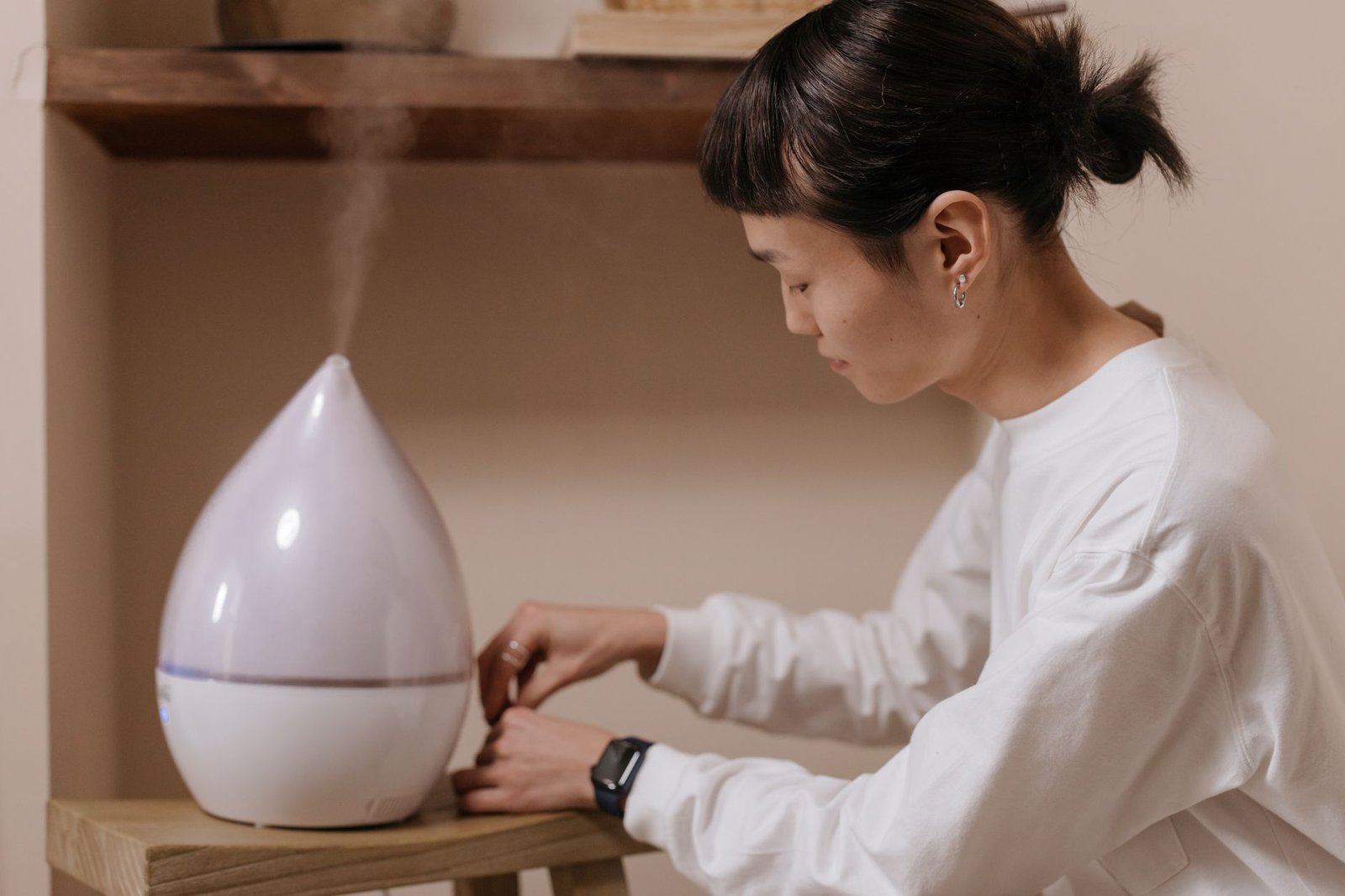 Person adjusting a white drop-shaped humidifier with a light mist rising, used to add moisture back to an over-dried room