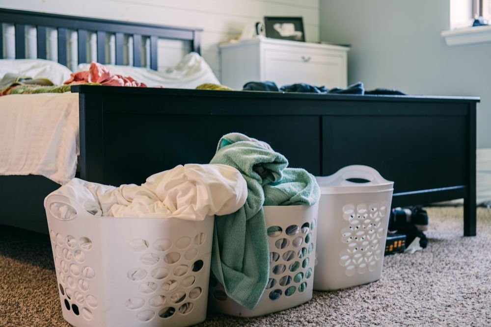 Two white laundry baskets of clothes and towels at the foot of a bed in a bedroom