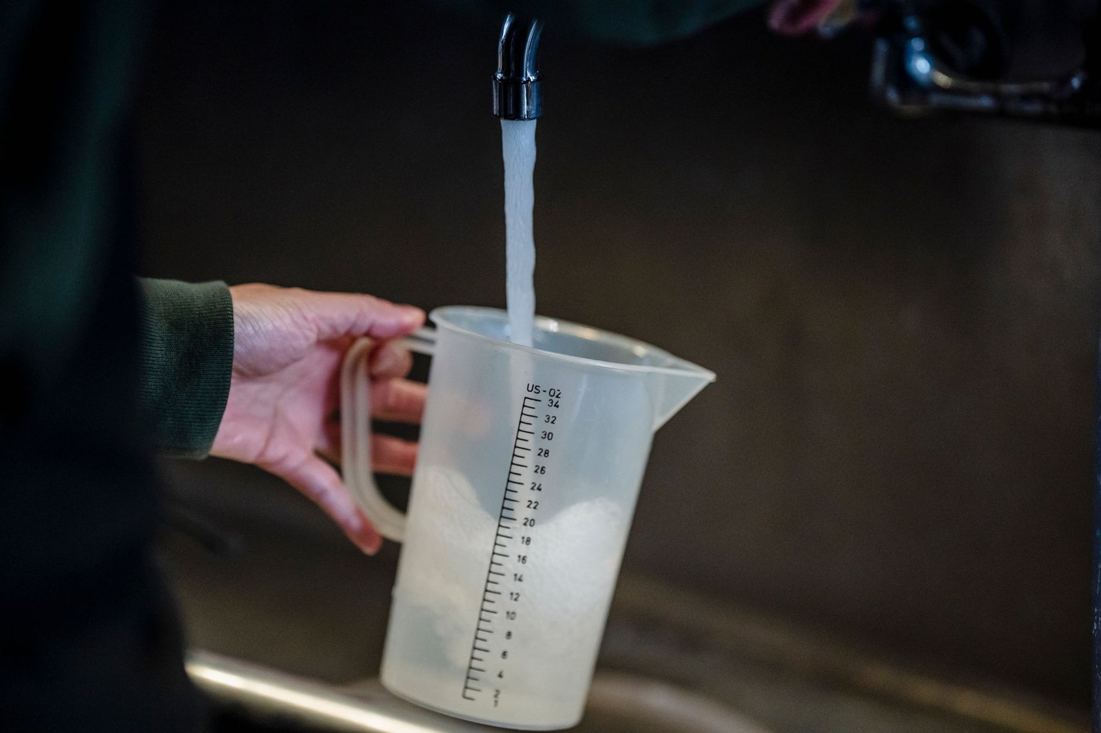 Hand holding a clear measuring jug under a running kitchen tap, the simplest way to measure how much water a dehumidifier collects