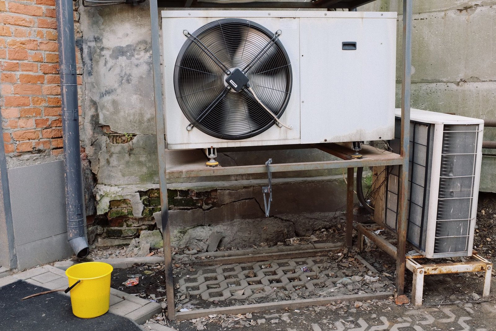 Two outdoor air conditioner condenser units, one with a large fan, mounted on a metal frame against a weathered wall
