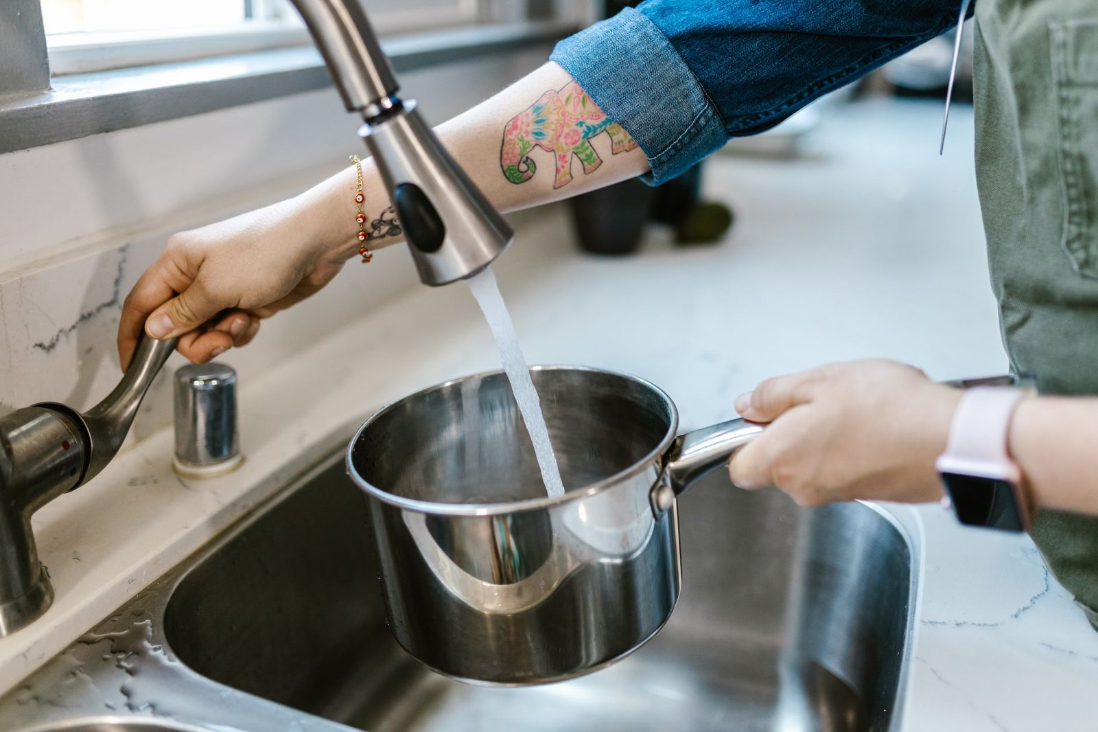 Hands holding a stainless steel pan under a running tap in a kitchen sink, the same sink where a dehumidifier tank is emptied and rinsed