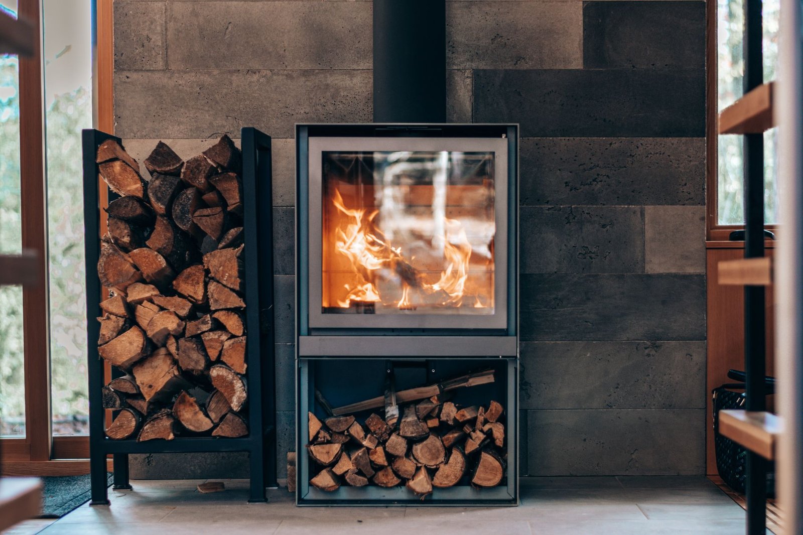 Modern wood stove with a glass door and neatly stacked split logs in a metal rack beside and below it