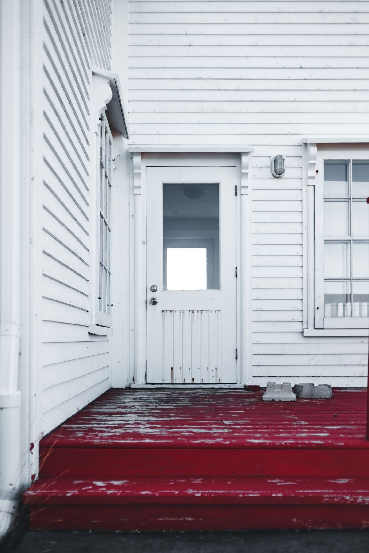 Timber porch step with worn red paint beside a white painted door and a downpipe at the corner