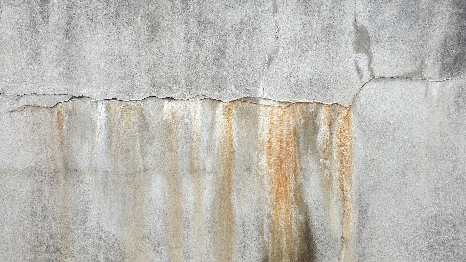 Grey concrete wall with cracks and rust-coloured water streaks running down from a horizontal crack