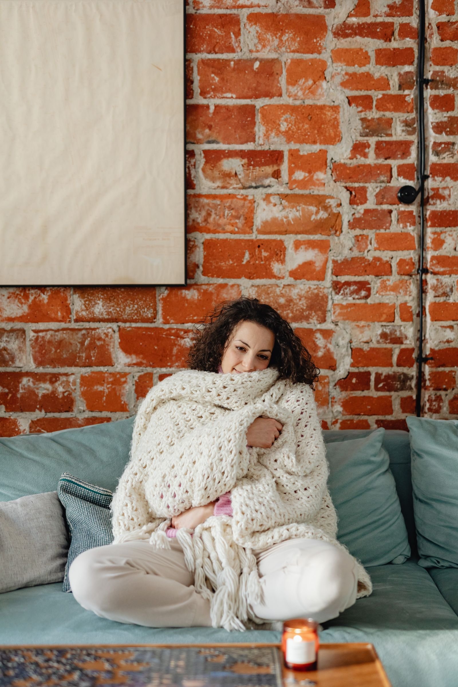 Woman wrapped in a chunky knit blanket on a sofa in front of a brick wall